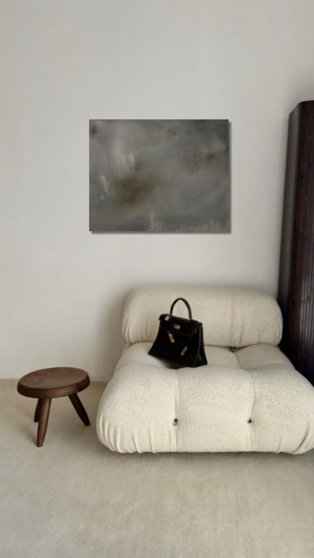 Beige armchair with a black bag on a light gray floor, wooden stool, and abstract painting on a white wall.